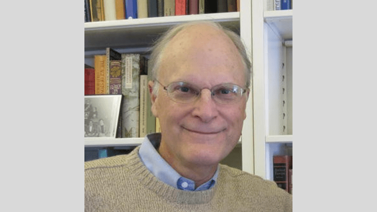 Professor Emeritus Timothy Earle is pictured in front of a bookshelf. He is wearing clear eyeglasses and a tan sweater over a blue collared shirt.