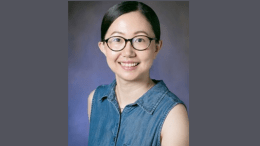 Professor Xiumin Du is pictured smiling in front of a dark gray background. She is wearing black eyeglasses and a blue collared shirt.